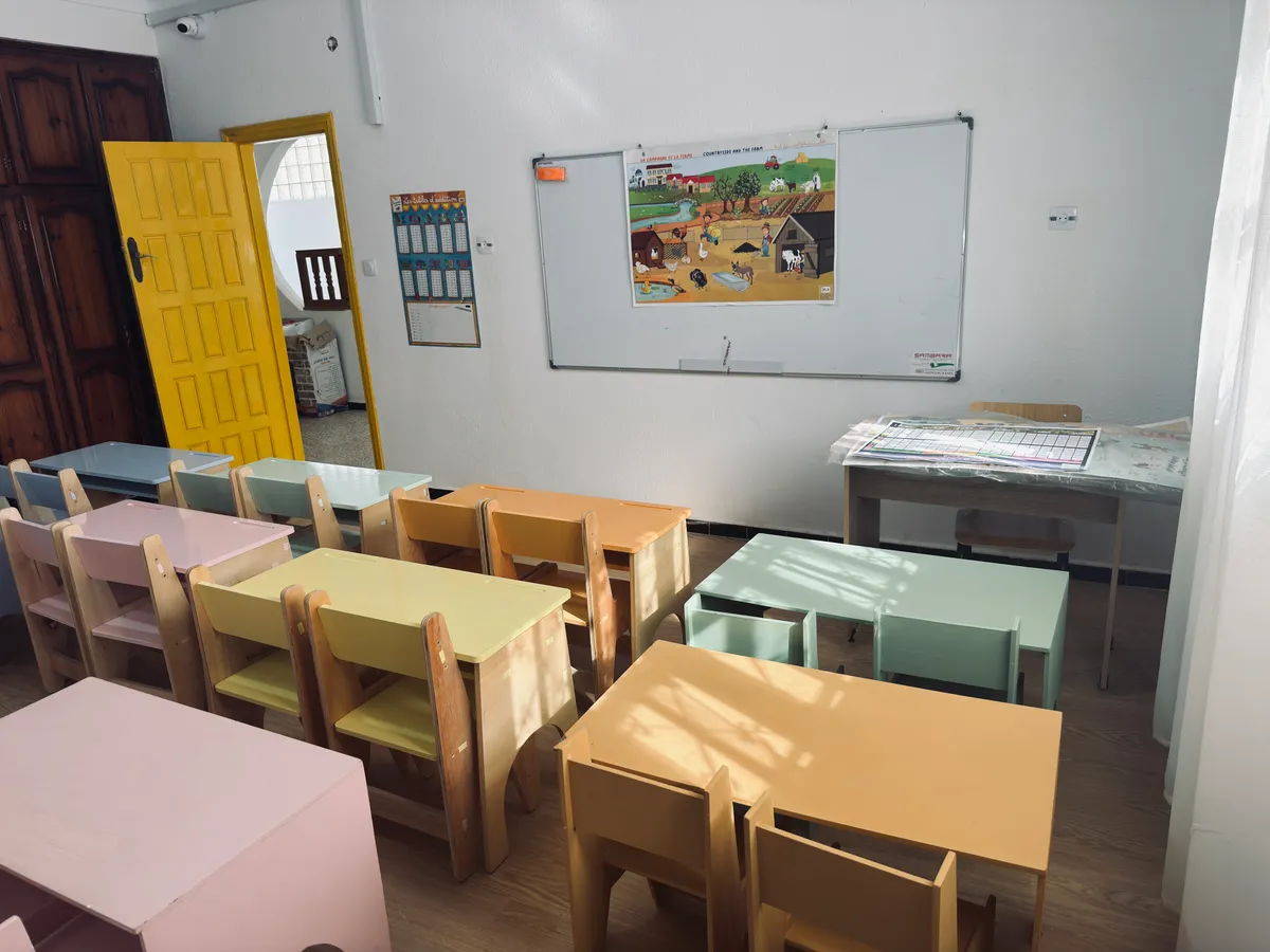 Classe aux petits bureaux pastel, porte jaune et tableau blanc avec une affiche de la ferme.