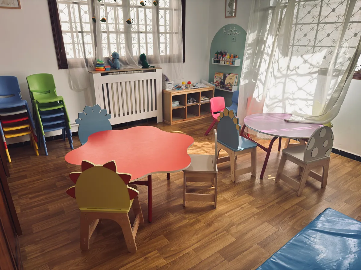 Salle d’activités avec table rouge en forme de fleur, chaises colorées et coin lecture.