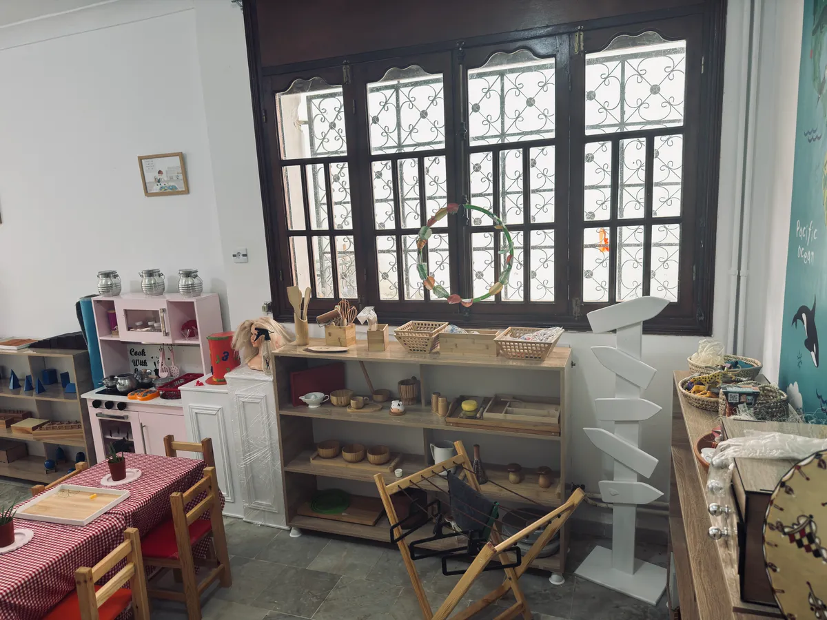 Étagères en bois sous une grande fenêtre, avec paniers, plateaux et objets du quotidien à hauteur d’enfant.