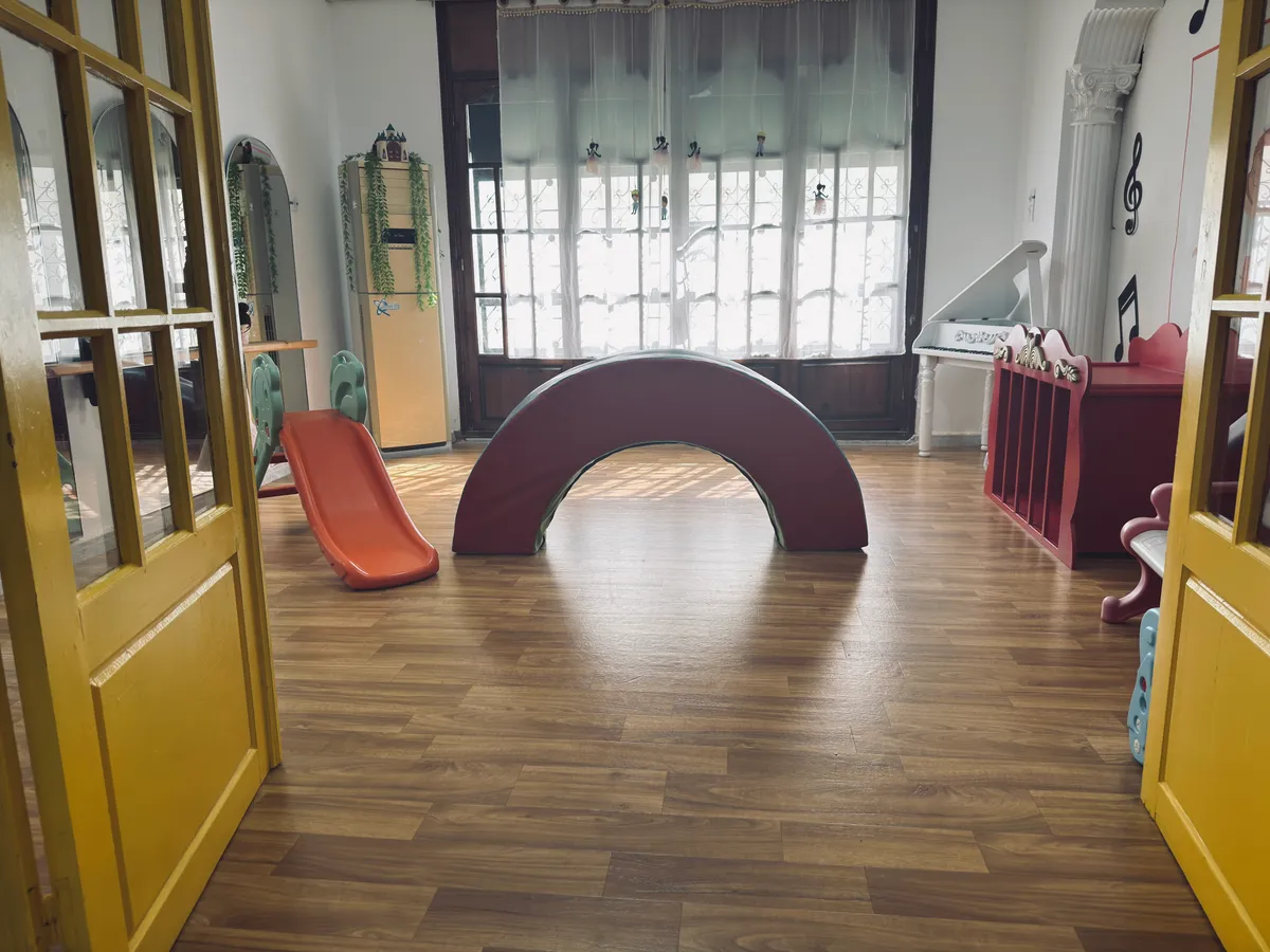 Salle de motricité au parquet clair : arche à bascule, petit toboggan et grandes fenêtres.