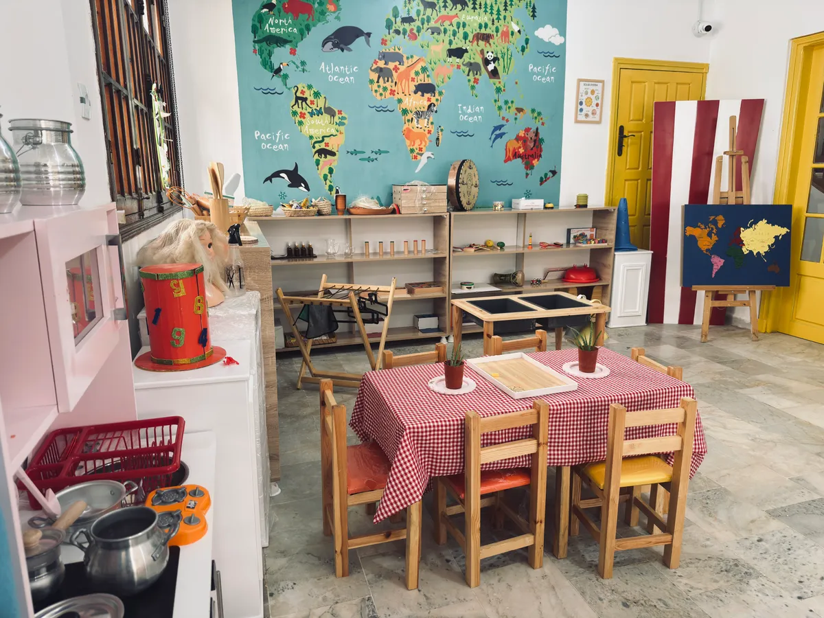 Salle Montessori : table à nappe vichy, coin cuisine pour enfants et grande carte du monde au mur.