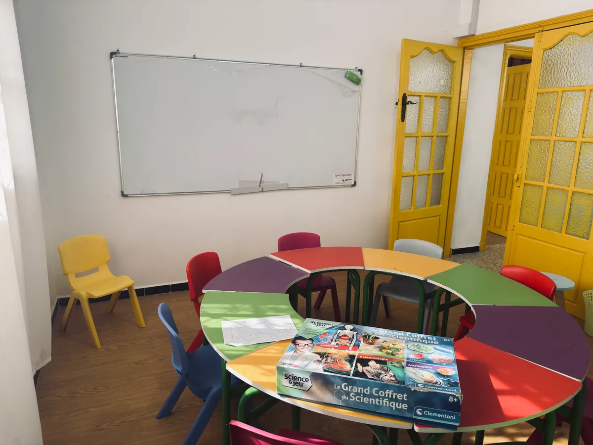 Table ronde colorée avec un coffret scientifique, chaises d’enfant et tableau blanc.