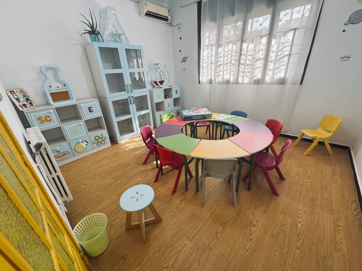 Salle des sciences : table ronde multicolore, meubles bleu clair et étagère en forme de fiole.