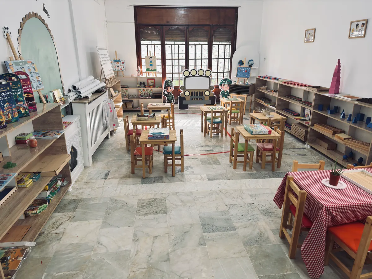 Grande salle Montessori lumineuse avec étagères basses, petites tables et chaises en bois.