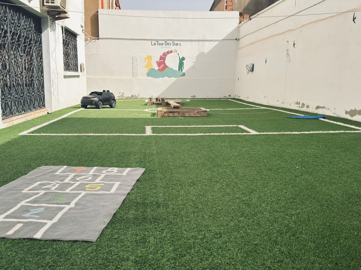 Cour extérieure en gazon synthétique avec marelle, petite voiture et fresque de la crèche au fond.