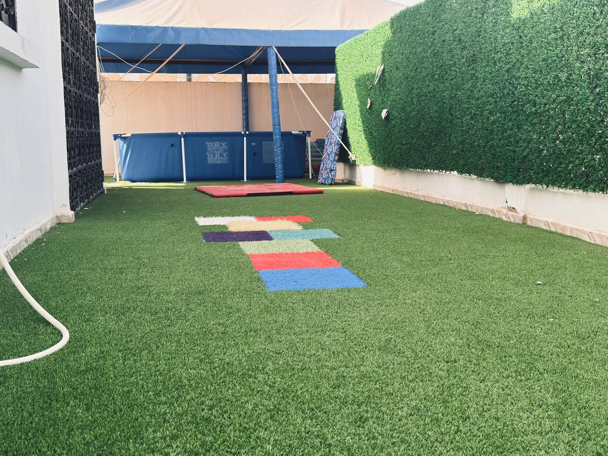 Terrasse en gazon synthétique avec dalles colorées, haie verte et espace couvert au fond.